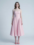 A woman models a pink and white gingham midi dress with a square neckline and a full, pleated skirt, standing against a plain background.