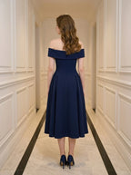 Back view of a woman in a navy blue off-the-shoulder midi dress with a pleated skirt, standing in a hallway.