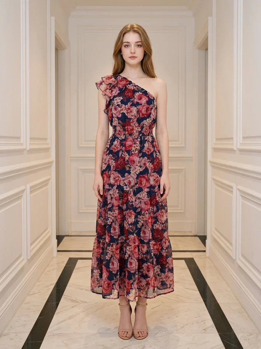 Woman wearing a navy floral print one-shoulder dress with a tiered skirt and ruffle detail, standing in a hallway.