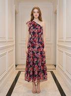 Woman wearing a navy floral print one-shoulder dress with a tiered skirt and ruffle detail, standing in a hallway.