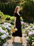 A woman walks through a garden wearing a black velvet midi dress with a floral print and carrying a straw basket.