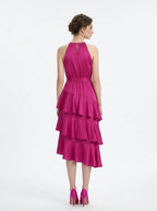 Back view of a woman in a fuchsia satin halter tiered midi dress with a self-tie belt.