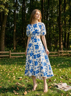 Woman wearing a white and blue floral print midi dress with cut-out waist and puff sleeves, standing in a grassy area with trees in the background.
