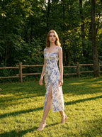 Woman wearing a floral print midi dress with tie shoulder straps and a high slit, walking in a grassy park.