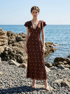 Woman wearing a brown polka dot midi dress with ruffled sleeves and a v-neckline, standing on a rocky beach.