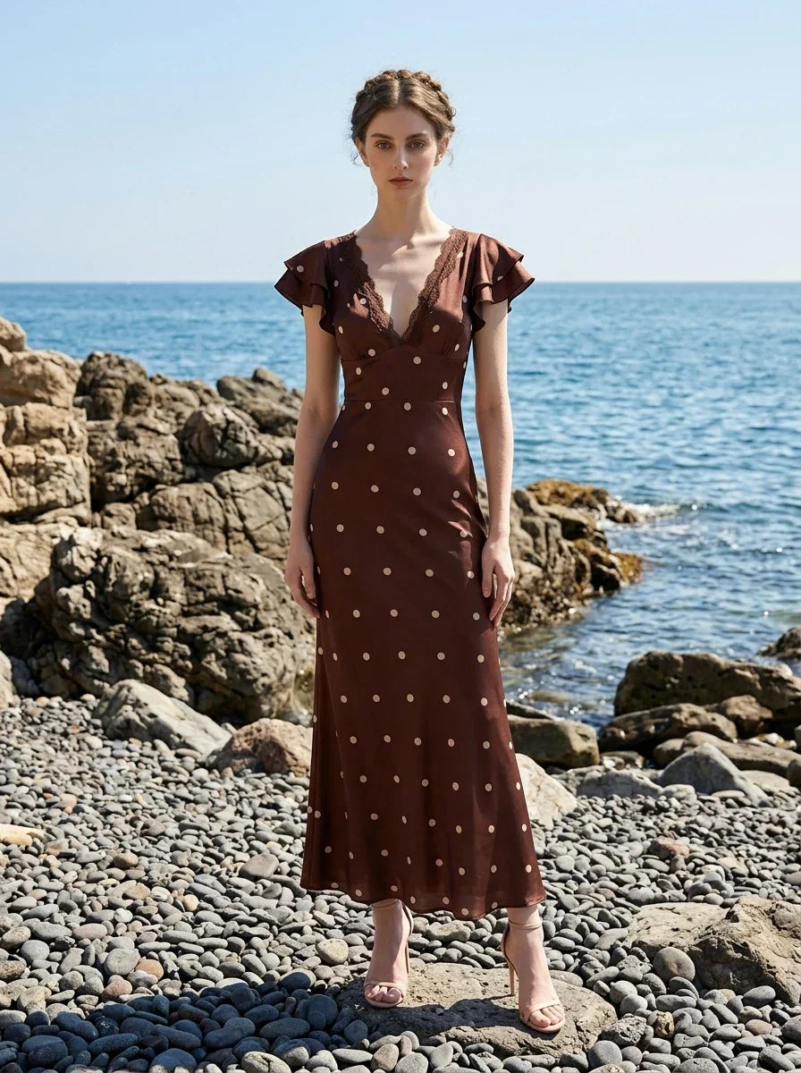 Woman wearing a brown polka dot midi dress with ruffled sleeves and a v-neckline, standing on a rocky beach.
