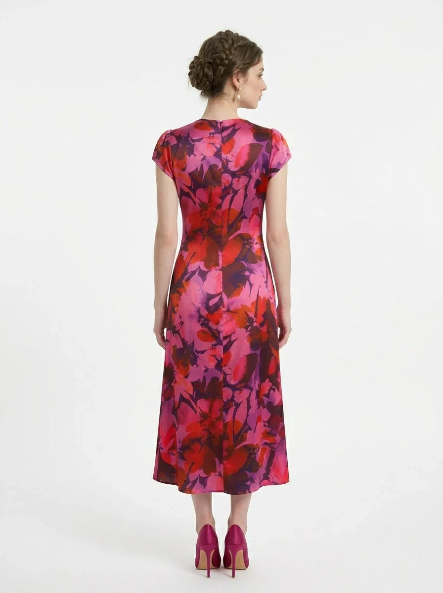 Back view of a woman wearing a pink and red floral print satin midi dress with short sleeves and ruched detailing, standing against a white background.