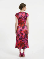 Back view of a woman wearing a pink and red floral print satin midi dress with short sleeves and ruched detailing, standing against a white background.