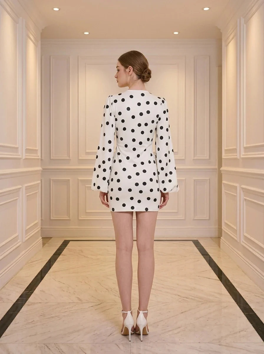 Back view of a woman standing in a white hallway wearing a white polka dot mini dress with long bishop sleeves and white heels.