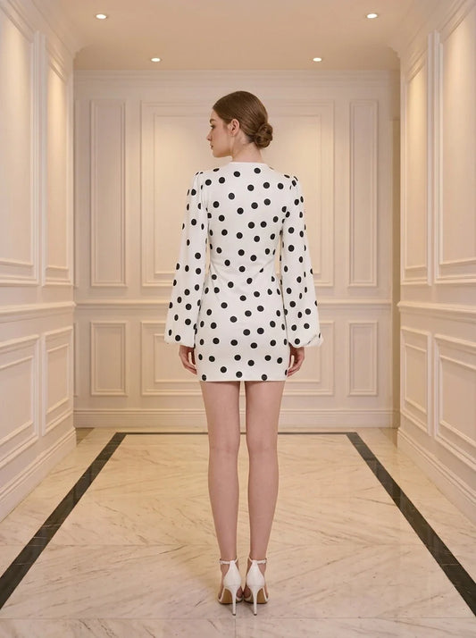 Back view of a woman standing in a white hallway wearing a white polka dot mini dress with long bishop sleeves and white heels.