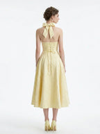 Back view of a woman in a yellow halter neck midi dress with corset detailing and a bow at the neck.