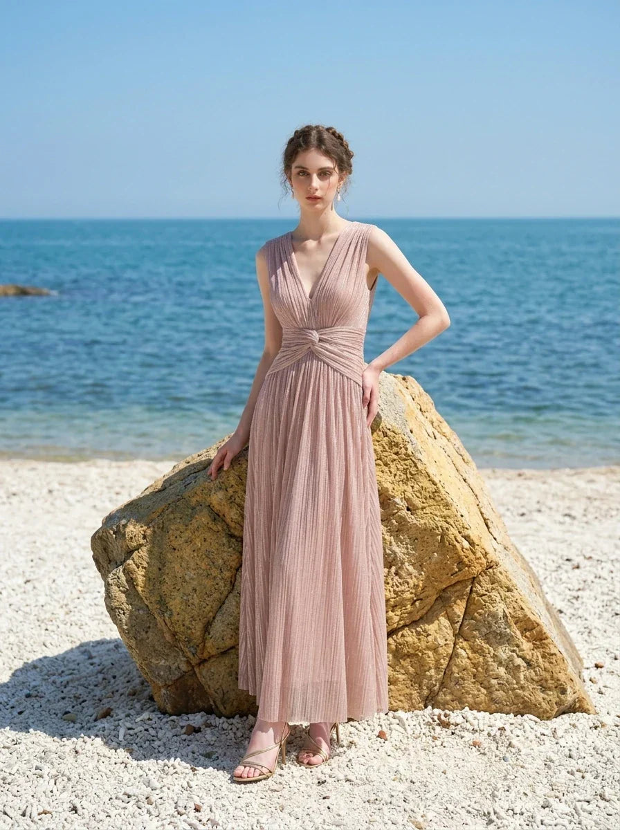 Woman in a rose gold shimmering metallic A-line V-neck sleeveless floor-length formal gown, standing by a rock on a beach
