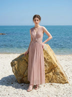 Woman in a rose gold shimmering metallic A-line V-neck sleeveless floor-length formal gown, standing by a rock on a beach