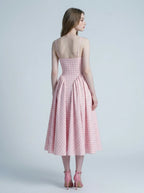 A woman models the back of a pink and white gingham midi dress with a square neckline and a full, pleated skirt, standing against a plain background.