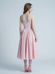 A woman models the back of a pink and white gingham midi dress with a square neckline and a full, pleated skirt, standing against a plain background.