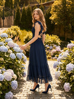 Woman walking in a garden pathway wearing a navy blue midi dress with ruffle sleeves and a tiered ruffled hem.