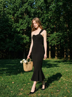 Woman in a black asymmetrical one-shoulder midi dress with a ruffled hem, carrying a straw bag, walking in a park with trees.