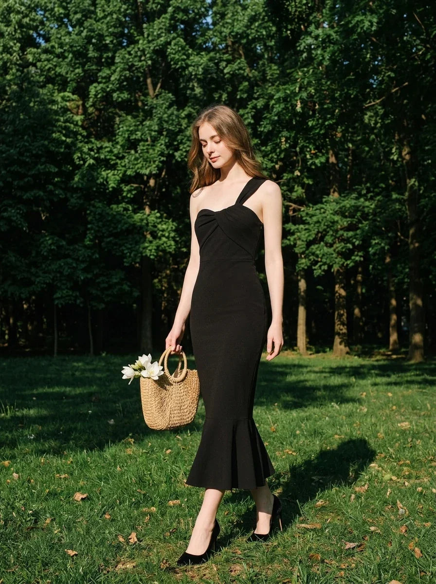 Woman in a black asymmetrical one-shoulder midi dress with a ruffled hem, carrying a straw bag, walking in a park with trees.