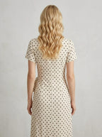 Back view of a woman wearing a cream and black polka dot midi wrap dress with short sleeves, standing against a white textured wall.