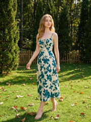 Woman wearing a floral print midi dress with spaghetti straps and a tie front detail, walking in a grassy park area with fallen leaves.