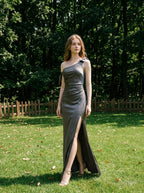 Woman wearing a floor-length grey satin one-shoulder dress with a high slit and bow accents, standing in a grassy field with trees in the background.