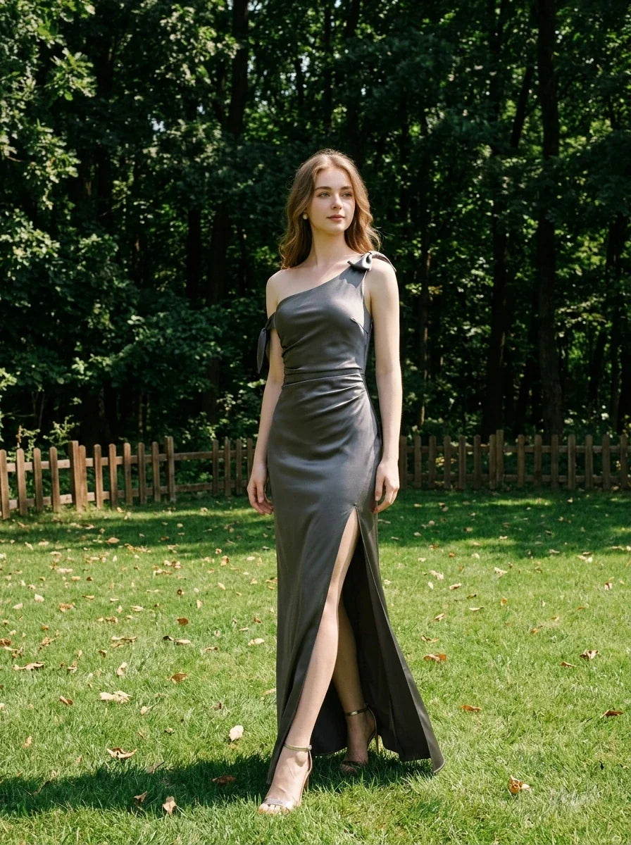 Woman wearing a floor-length grey satin one-shoulder dress with a high slit and bow accents, standing in a grassy field with trees in the background.