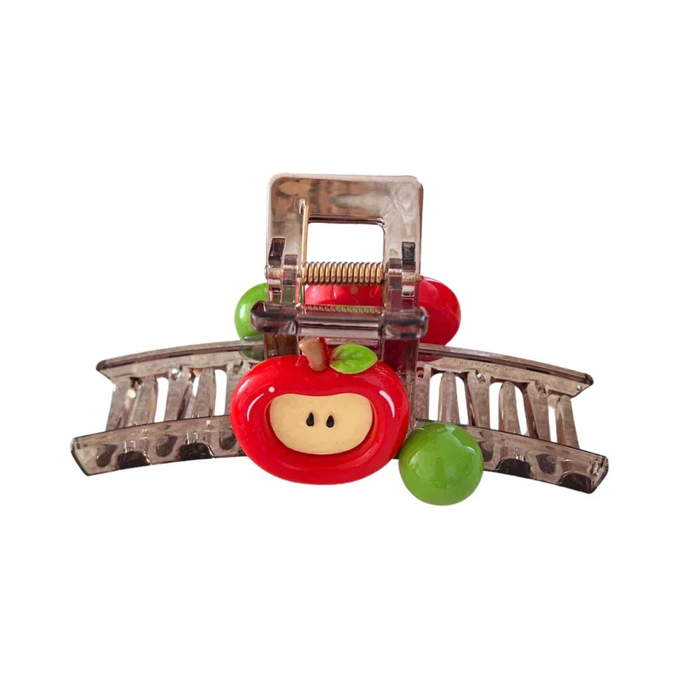 A close-up of a transparent hair claw clip with a red apple charm and green beads, against a white background. Main image