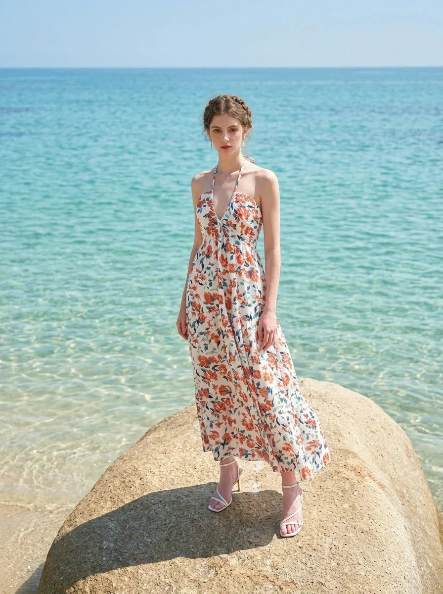 Woman wearing a white floral print maxi dress with a halter neckline and tiered skirt, standing on a rock in the ocean.
