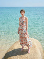 Woman wearing a white floral print maxi dress with a halter neckline and tiered skirt, standing on a rock in the ocean.