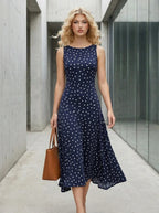 Woman wearing a navy blue sleeveless midi dress with white polka dots, walking with a brown tote bag.