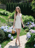 Woman wearing a white floral print mini dress with spaghetti straps and a sweetheart neckline, carrying a woven basket, walking on a garden path surrounded by hydrangeas.