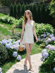 Woman wearing a white floral print mini dress with spaghetti straps and a sweetheart neckline, carrying a woven basket, walking on a garden path surrounded by hydrangeas.