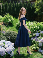 Woman wearing a navy blue off-the-shoulder midi dress with a pleated skirt, turning to look over her shoulder in a garden.