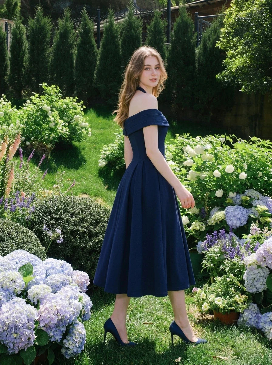 Woman wearing a navy blue off-the-shoulder midi dress with a pleated skirt, turning to look over her shoulder in a garden.