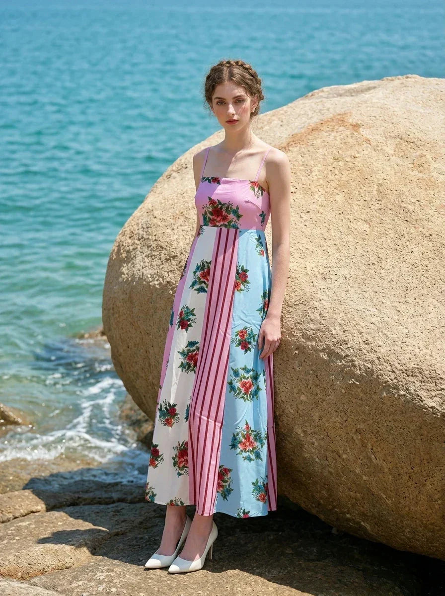 woman in a pink, blue, and white floral print maxi dress with spaghetti straps, leaning against a large rock on a beach with the ocean in the background