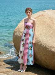 woman in a pink, blue, and white floral print maxi dress with spaghetti straps, leaning against a large rock on a beach with the ocean in the background