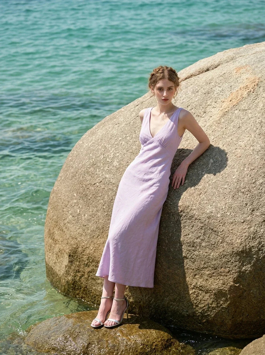 Model wearing a lavender midi dress with a V-neckline and sleeveless design, leaning against a large rock in the ocean.