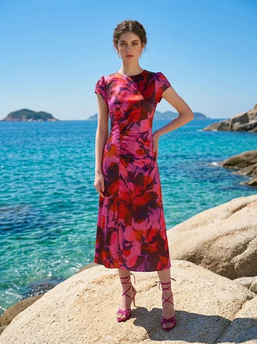 Woman wearing a pink and red floral print satin midi dress with short sleeves and ruched detailing, standing on rocks on a beach with the ocean in the background.