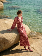 woman wearing a pink and red floral print satin maxi wrap dress with long bishop sleeves and a v-neck, sitting on a large rock on a beach with the ocean in the background