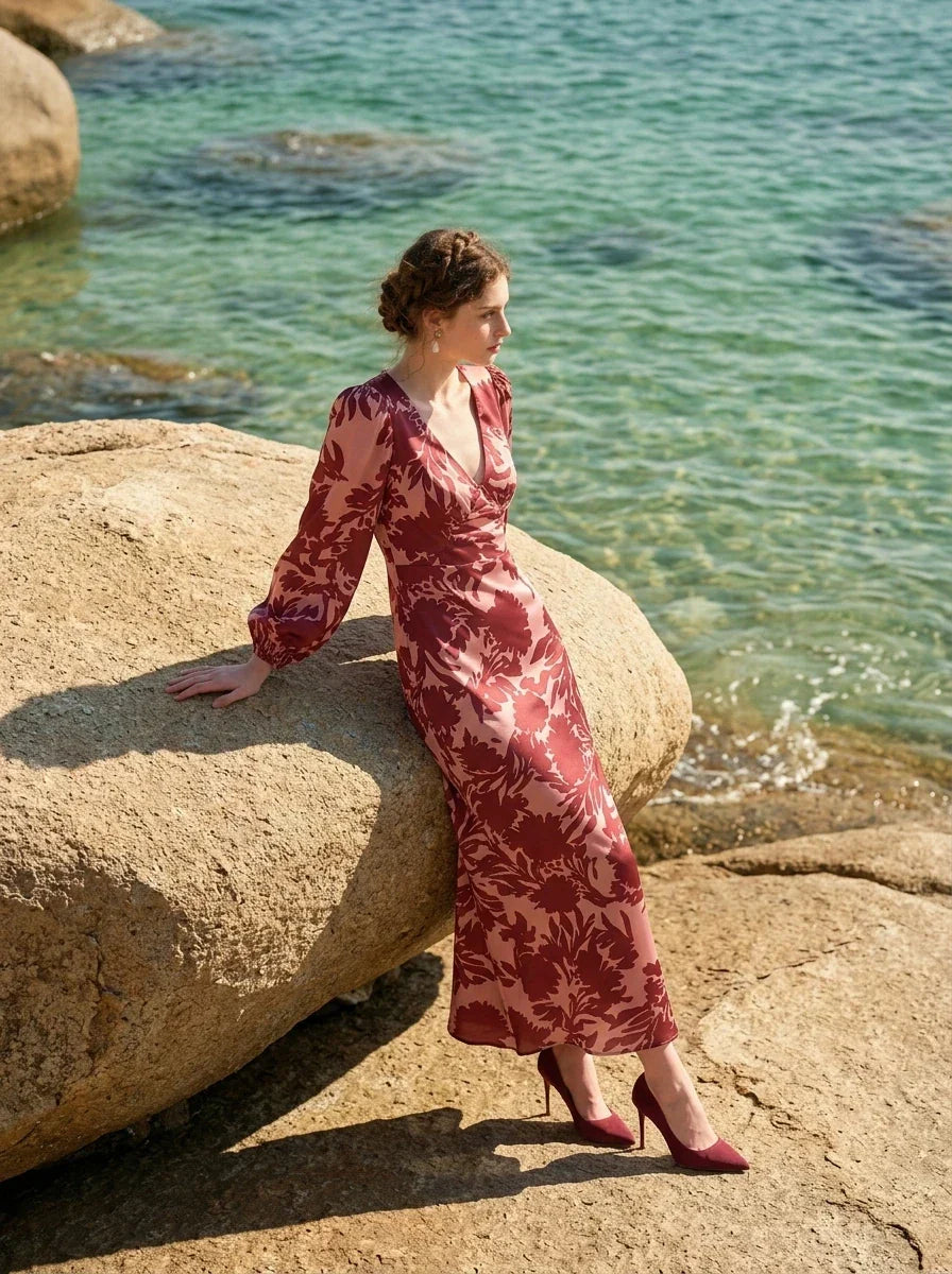 woman wearing a pink and red floral print satin maxi wrap dress with long bishop sleeves and a v-neck, sitting on a large rock on a beach with the ocean in the background