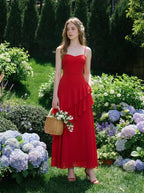 Woman wearing a red floor-length chiffon dress with tiered ruffles and spaghetti straps, holding a wicker basket with flowers in a garden setting.