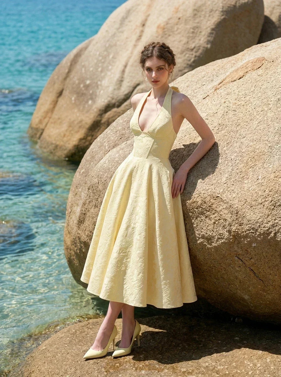 Woman wearing a yellow halter neck midi dress with a full skirt, standing near large rocks by the sea.