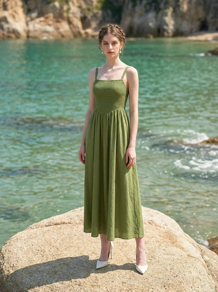 Woman wearing a green linen midi dress with a smocked bodice and square neckline, standing on a large rock in the ocean.