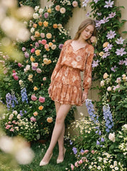 Woman leaning against a wall covered in flowers, wearing a floral print mini dress with long sleeves and a V-neck.