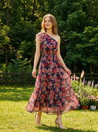 Woman wearing a navy floral print one-shoulder dress with a tiered skirt and ruffle detail, standing in a sunny garden.