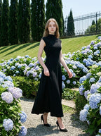 Woman in a black mock neck cap sleeve A-line dress walking along a path lined with hydrangeas.