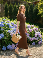 Woman walking in a garden wearing a brown ribbed knit dress with white trim on the collar and sleeves, holding a straw bag with flowers, and wearing nude high heels.