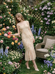 Woman wearing a cream-colored sleeveless midi dress with floral embellishments and a ruffled hem, standing in a garden with roses.