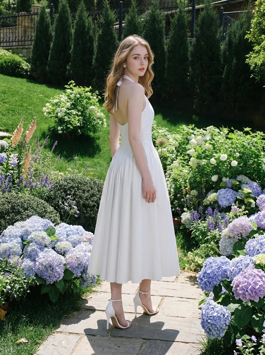 Woman in a white halter neck midi dress with an open back, standing in a garden with hydrangeas and other blooming flowers.