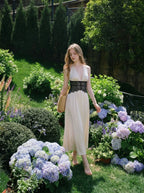 Woman in a cream satin maxi dress with black lace waist detail, walking through a garden with hydrangeas.
